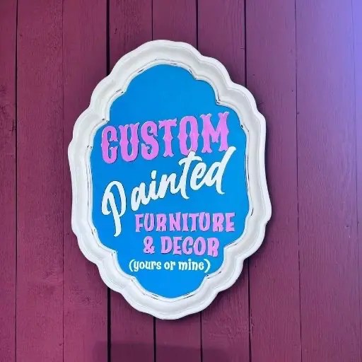 Blue and white vintage sign reading “Custom Painted Furniture & Decor (yours or mine)” on a purple wall at a painted furniture store near me.