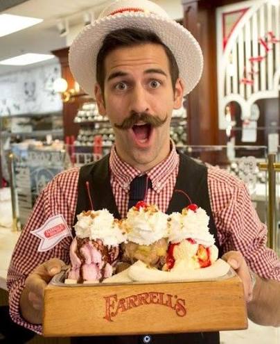 A smiling server wearing a bow tie and hat, holding a wooden tray filled with various colorful ice cream sundaes at Farrell's ice cream parlor.