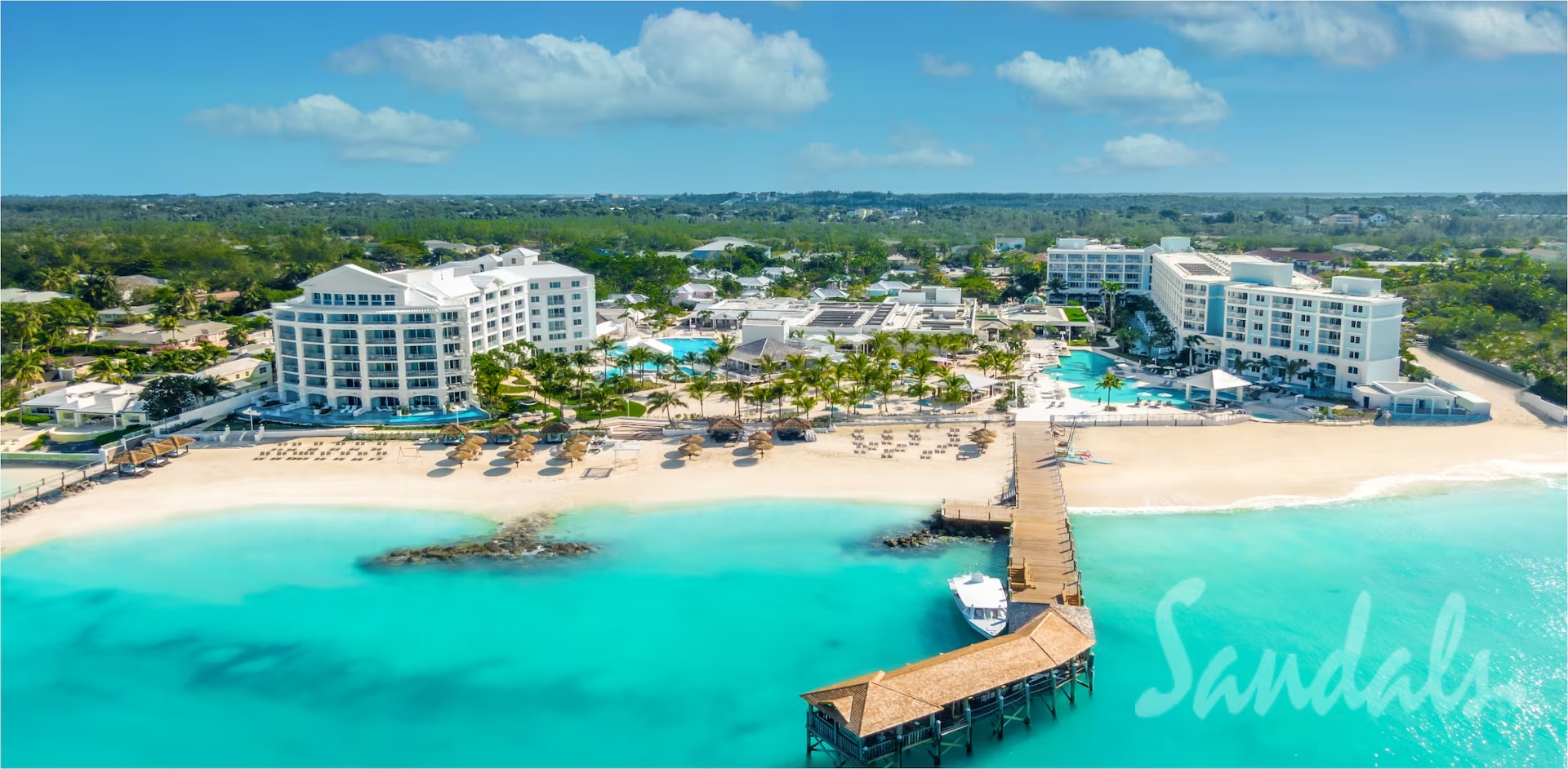Aerial view of a white resort with palm trees, a sandy beach, and turquoise ocean; a wooden pier extends into the sea.