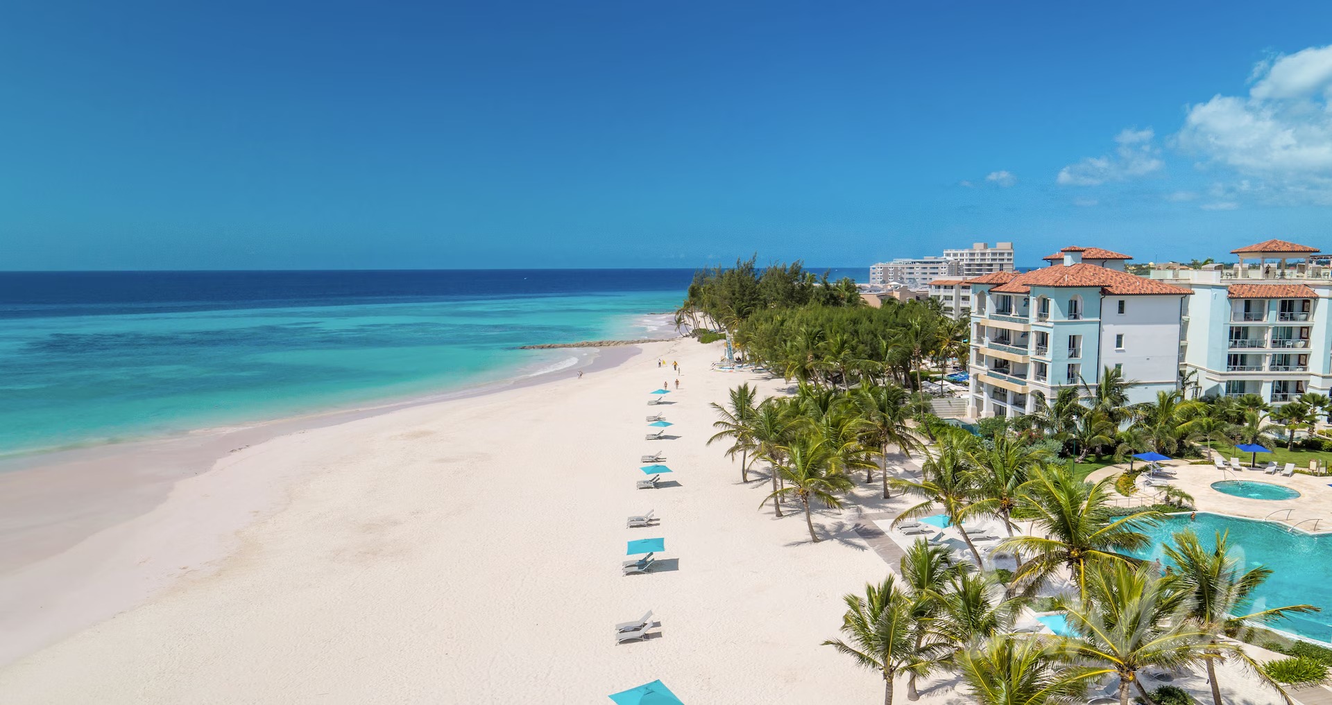 A tropical beach resort with white sand, turquoise water, palm trees, and buildings with red-tiled roofs nearby.