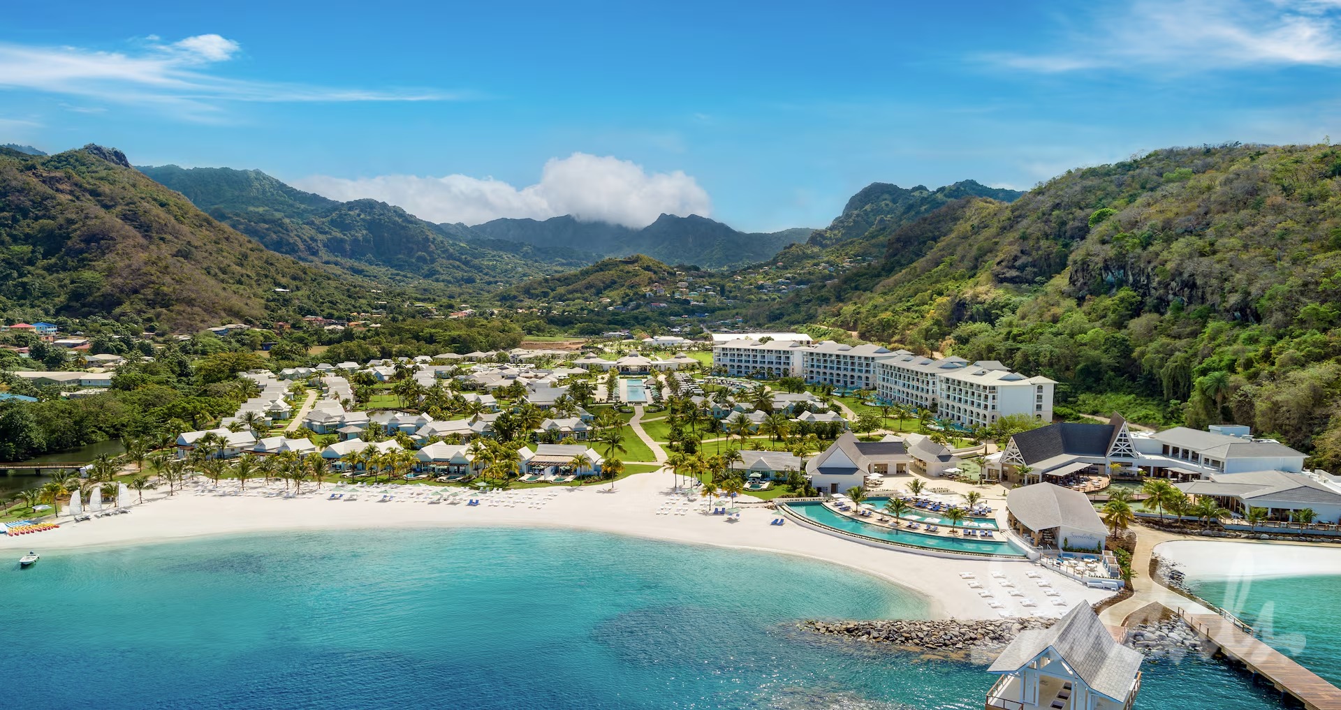 Tropical beach resort with white sand, turquoise water, palm trees, and green, mountainous backdrop.