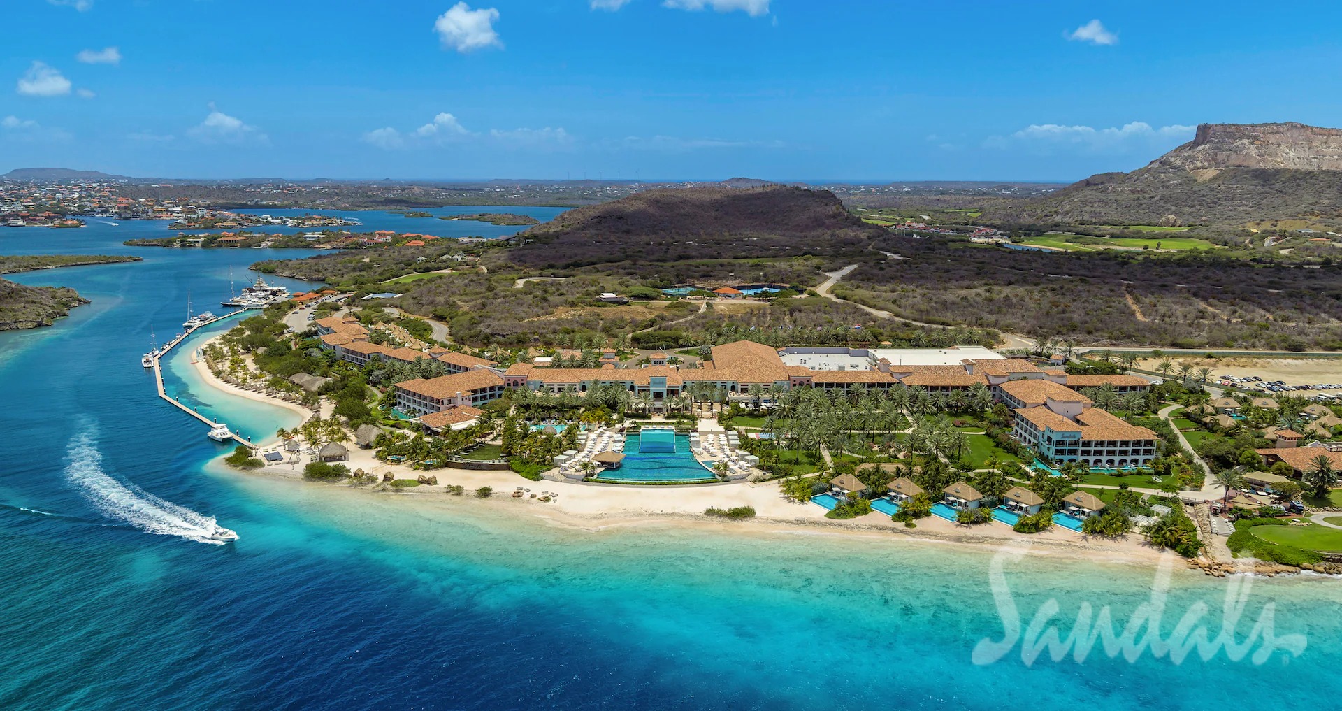 Aerial view of a tropical resort along a turquoise coastline with a large pool, palm trees, and sandy beaches.