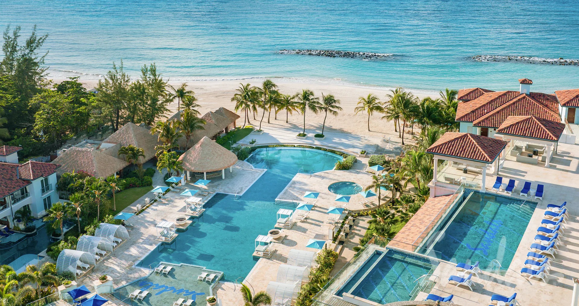Beachfront resort with a large pool, palm trees, and red-roofed buildings overlooking the ocean.