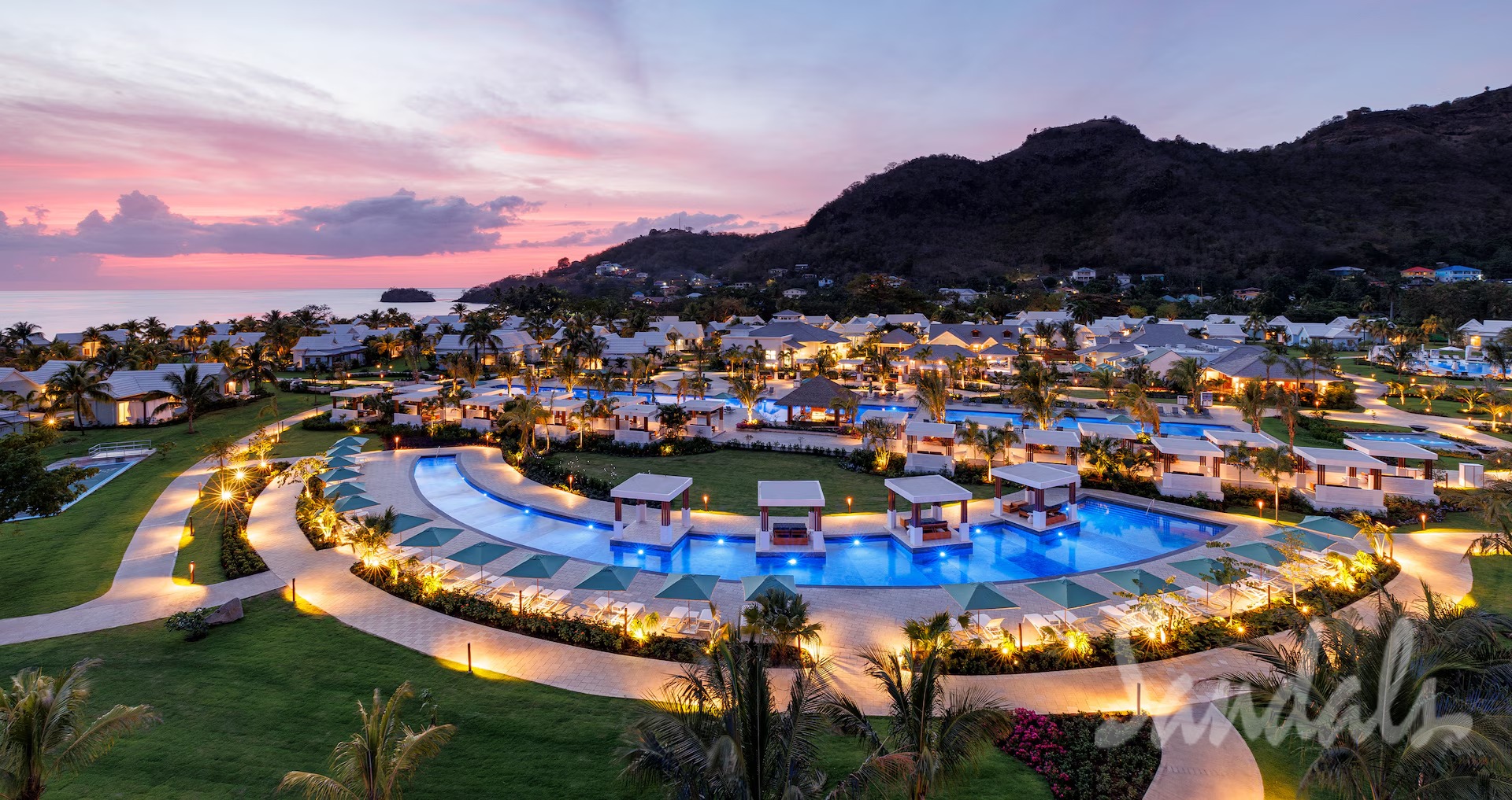 Aerial view of a tropical resort at sunset with illuminated pools, palm trees, and white villas against a mountainous backdrop by the coast.