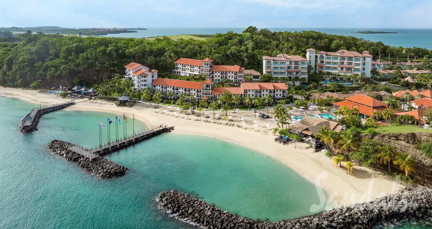 Aerial view of a tropical resort with red-roofed buildings, palm trees, and a curved white-sand beach beside turquoise water and a wooden pier.