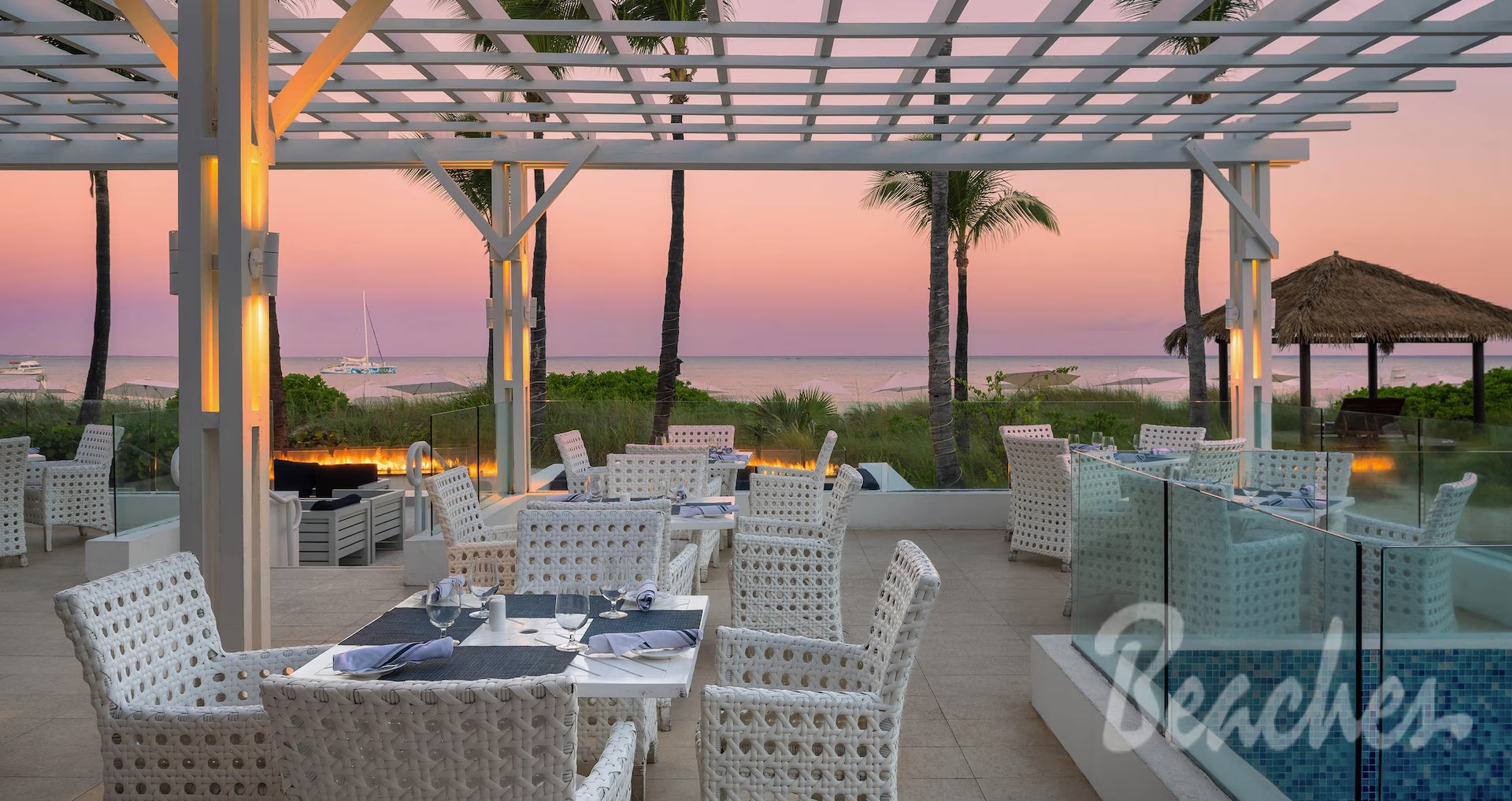 Outdoor seaside restaurant terrace at sunset with white wicker chairs and tables set for dinner overlooking the ocean and palm trees.