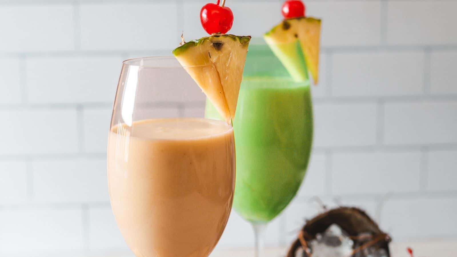 Two colorful smoothies in clear glasses, one orange and one green, each garnished with a pineapple slice and cherry, displayed against a tiled backsplash.