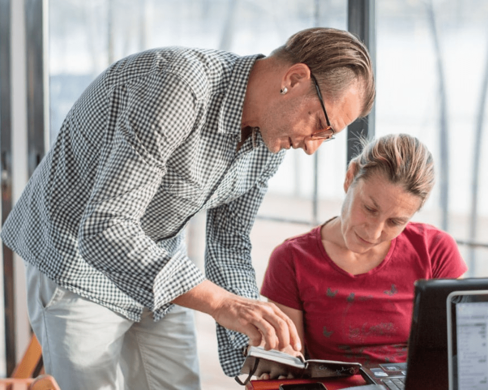 Two team members from Myriad Solutions a man and a woman discussing some notes in a notebook, one is sitting in front of a desk the other is standing over her