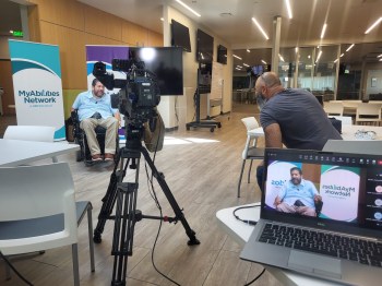 Humberto “Beto” Gurmilan, who is seated in a wheelchair, is filmed by a man seated next to a camera. The resulting shot of Gurmilan is seen on an open laptop.
