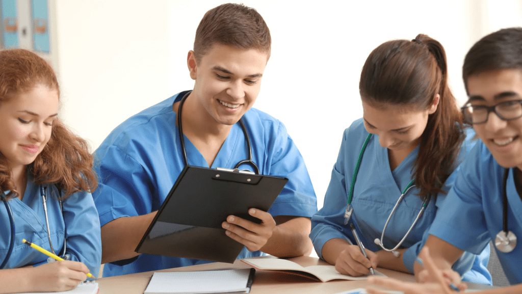 Four medical students in blue scrubs engaged in a collaborative study session, one holding a clipboard while others take notes.