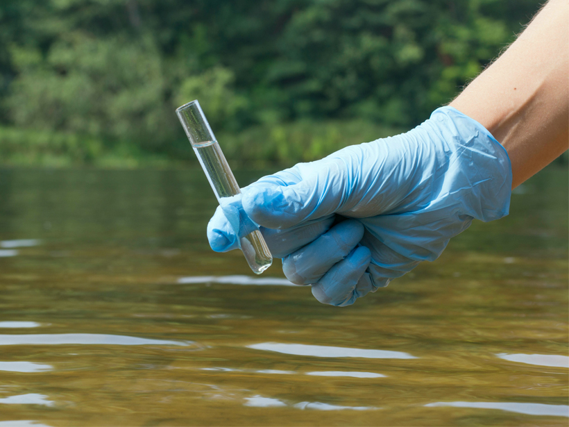 A gloved hand holds a test tube of water above a flowing river.