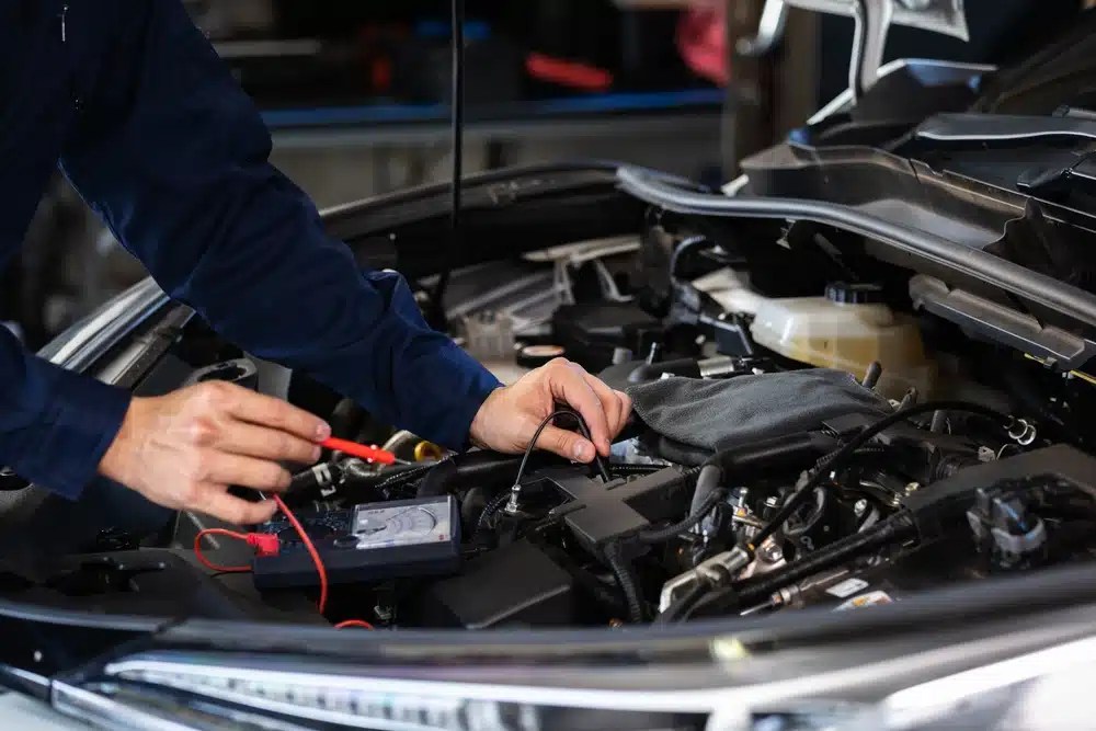 Mechanic in blue work shirt using a multimeter to test car battery electrical connections under open hood