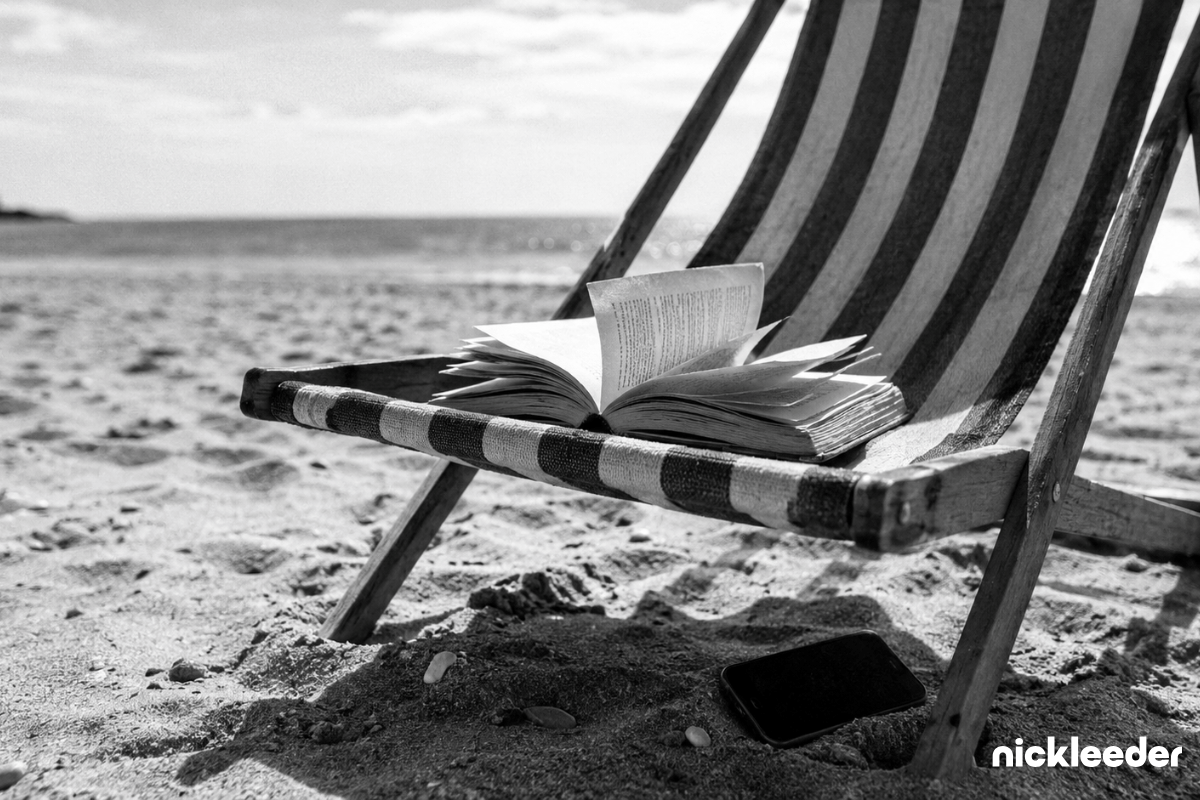 A paperback and a mobile phone left face down on a striped deckchair on a beach, nobody in sight.