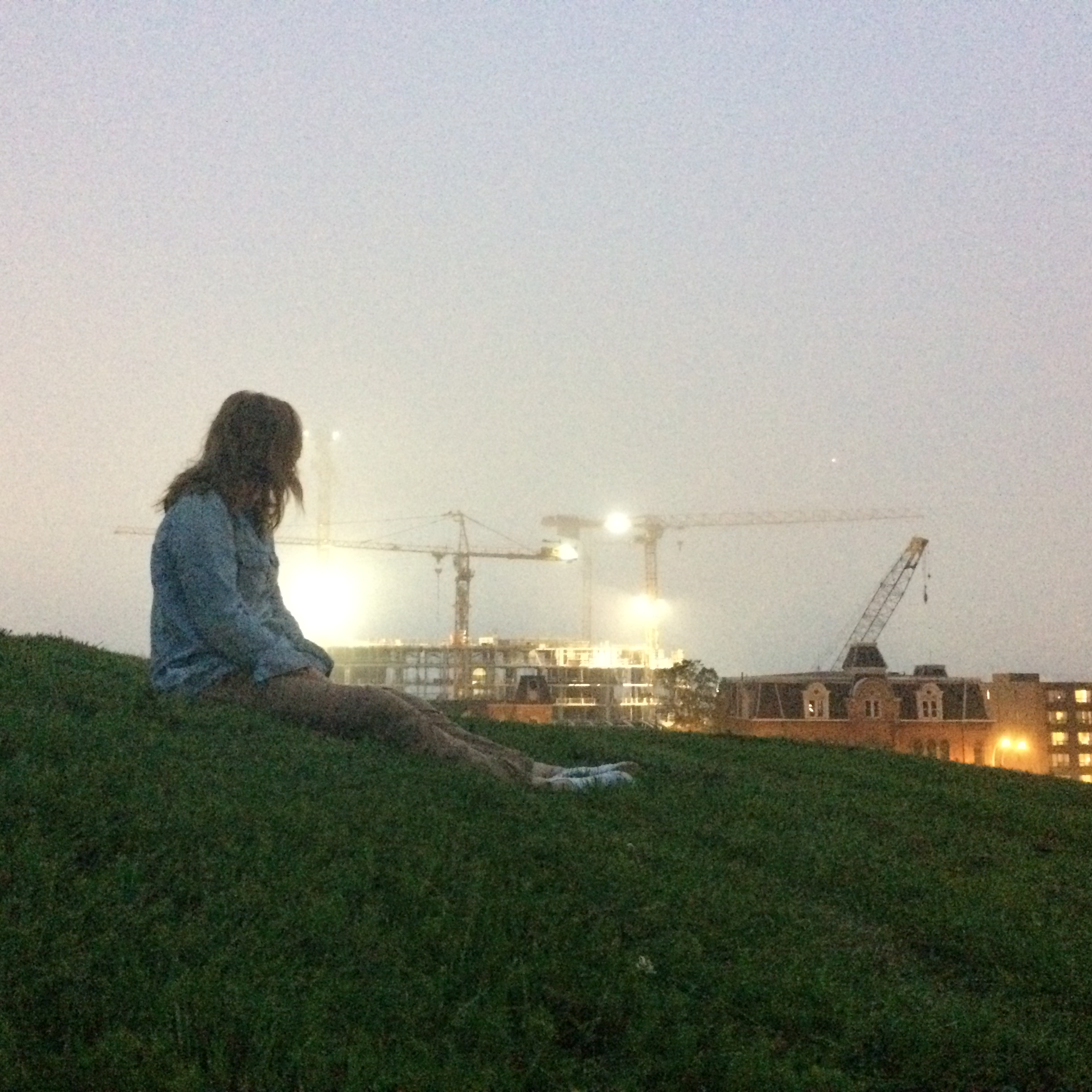 Photo of a woman sitting on the grass at dusk looking away from the camera at some construction.