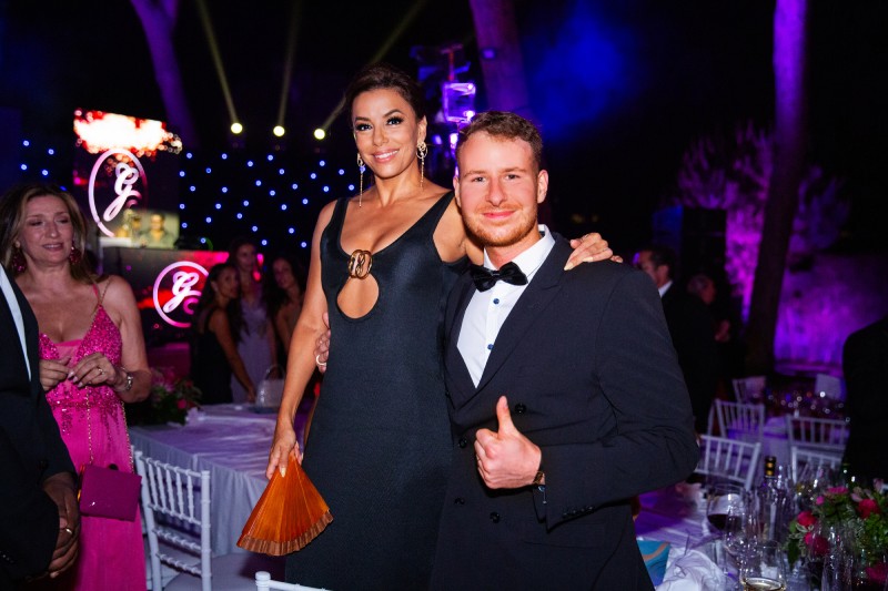 A woman in a stylish black dress with an elegant design poses confidently with a man in a tuxedo at a festive event, surrounded by onlookers and colorful lights.