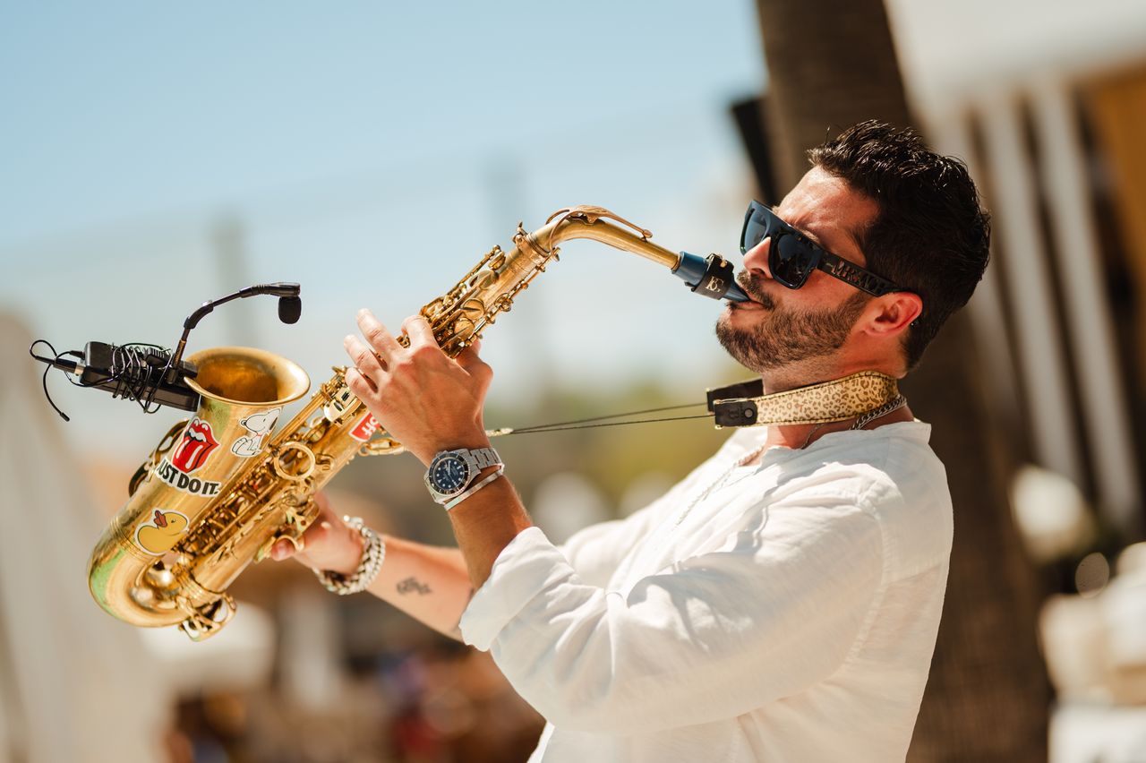 Un musicien joue du saxophone en plein air, vêtu d'une chemise blanche et de lunettes de soleil, dans un environnement festif de Nikki Beach Marbella.