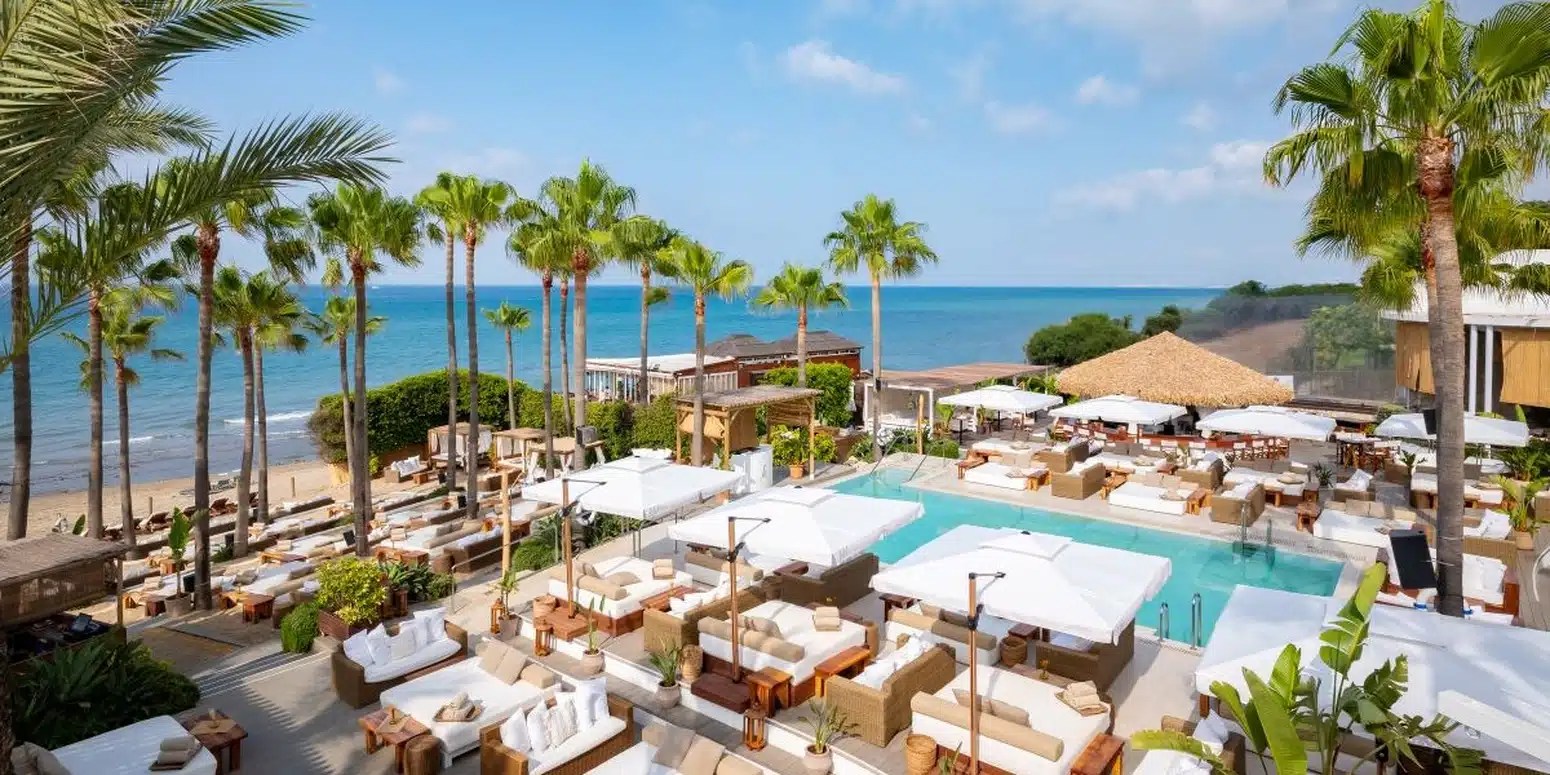 Vue panoramique de Nikki Beach Marbella, montrant la piscine au centre, des lits balinais élégants et la plage méditerranéenne en arrière-plan, entourée de palmiers sous un ciel ensoleillé.
