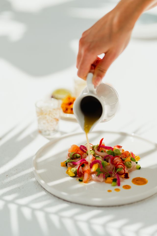 Une main verse une sauce sur un plat coloré de poisson cru, garnie de légumes frais et d'herbes, avec des verres à proximité.