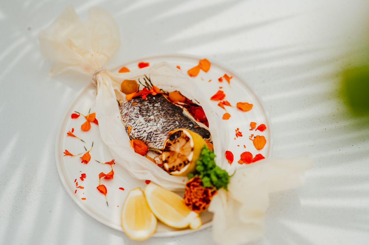 Un plat délicat de poisson cuit dans un papier, accompagné de tranches de citron et de pétales de fleurs comestibles, présenté sur une assiette élégante.