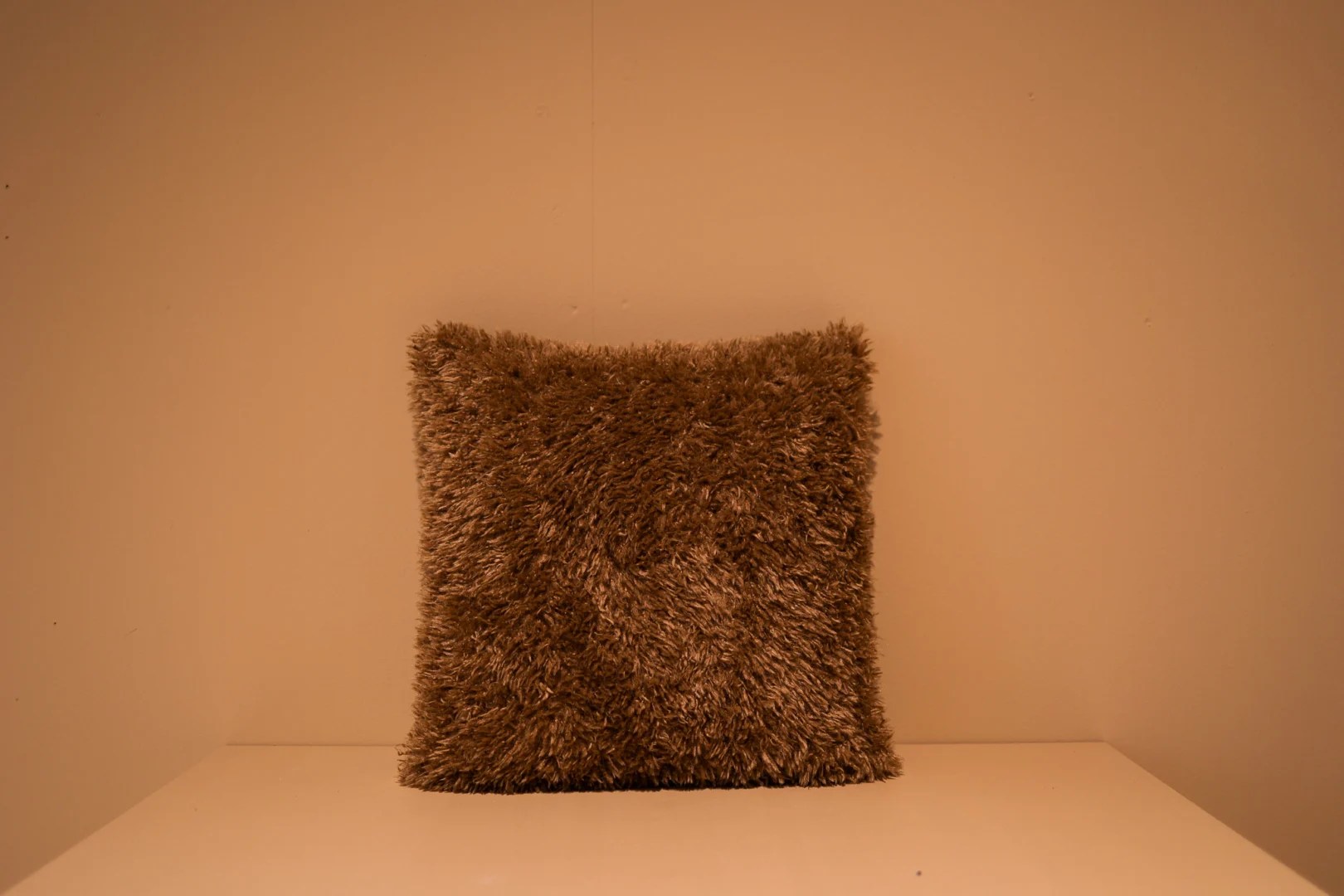 Het Armkussen 50x50, een vierkant kussen met bruine shaggy, staat rechtop tegen een beige muur op een wit vlak, verlicht met warm licht.