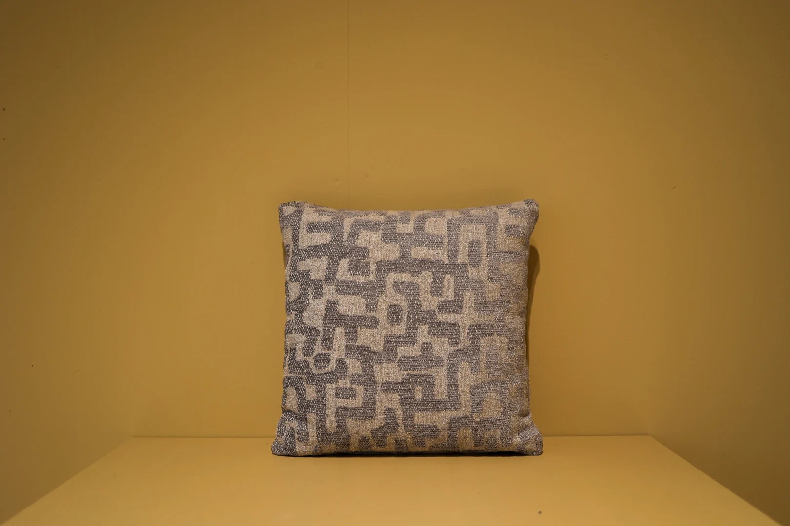 Het Armkussen 50x50, met een abstract grijs en beige geometrisch patroon met textuur, wordt getoond tegen een effen mosterdgele muur en oppervlak.