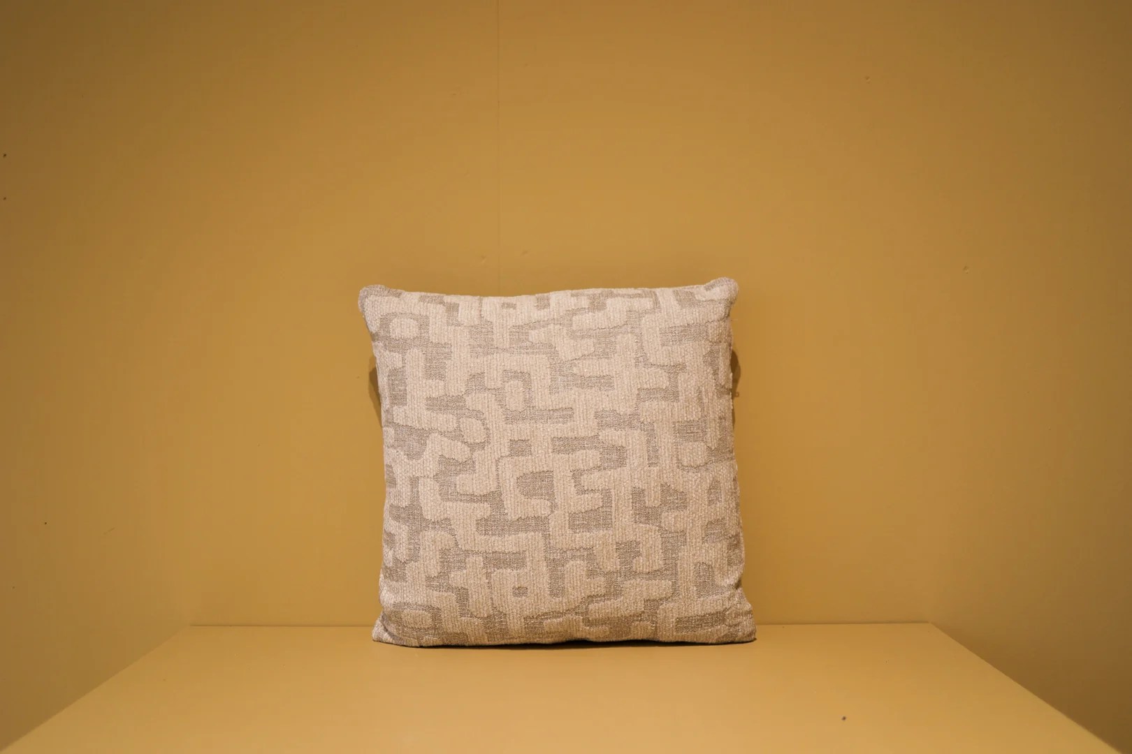 Het Armkussen 50x50, een vierkant kussen met een geometrisch patroon in beige en lichtgrijs, is gecentreerd tegen een effen bruine achtergrond en oppervlak.