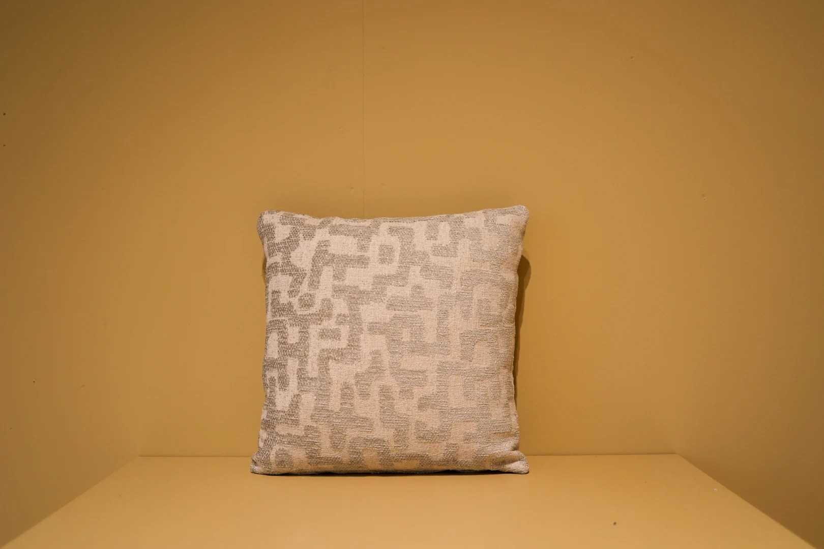Het Armkussen 50x50, een vierkant kussen met een lichtbeige en wit geometrisch patroon, ligt tegen een mosterdgele muur op een bijpassend geel vlak.