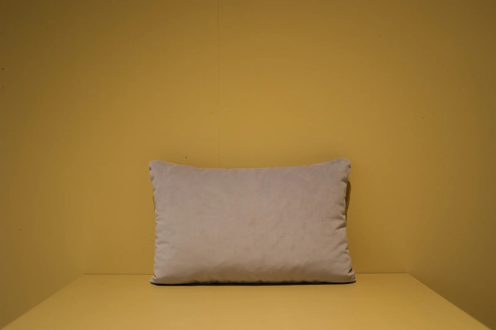 Het Armkussen 50x50, een effen lichtbeige rechthoekig kussen, rust tegen een effen mosterdgele muur en ondergrond, waardoor een eenvoudig en minimalistisch tafereel ontstaat.