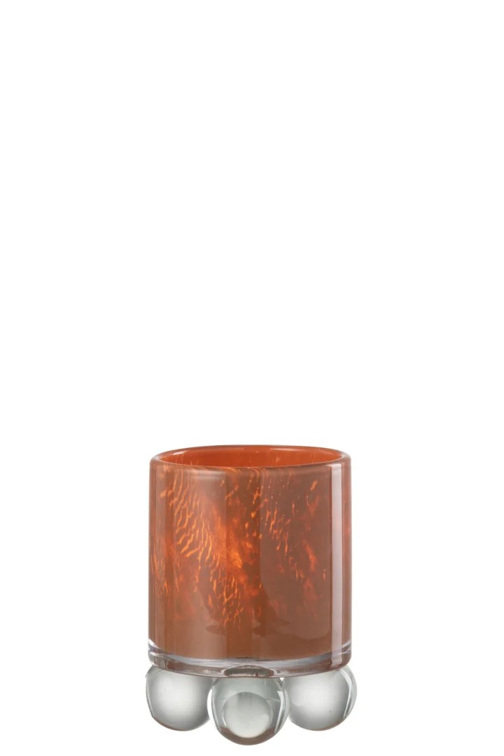 De Theelichthouder Glas Bruin Op Ballen is een korte glazen beker met een gemarmerd oranje patroon. Hij staat op een heldere voet met drie ronde, bubbelachtige glazen pootjes, afgebeeld tegen een witte achtergrond.