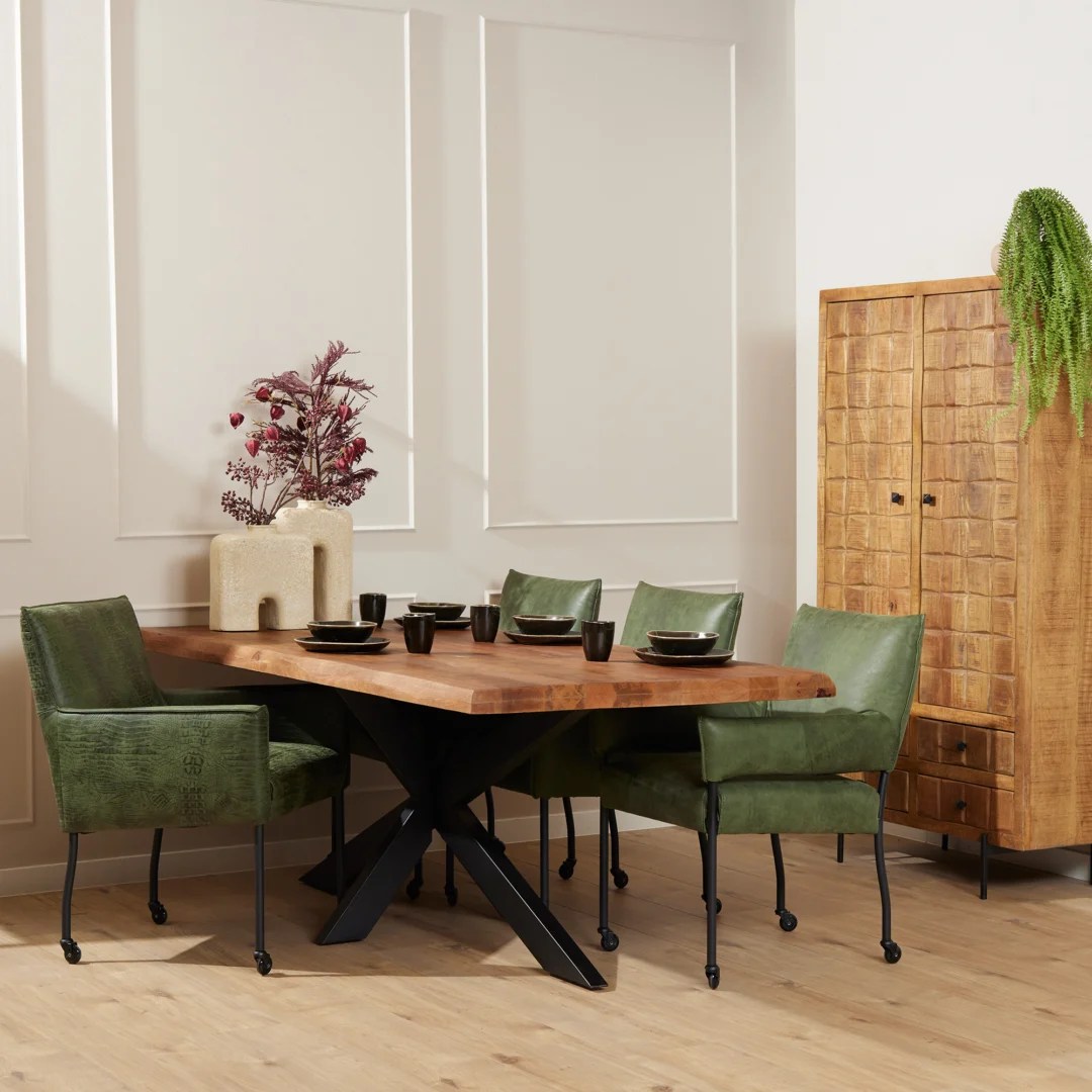 In een moderne eetkamer staat de Boomstamtafel Boston 180 cm met een zwart metalen onderstel, omringd door vier groen gestoffeerde stoelen. De tafel is gedekt met donker servies en een vaas met rode takken, met een hoge houten kast en een hangplant op de achtergrond.
