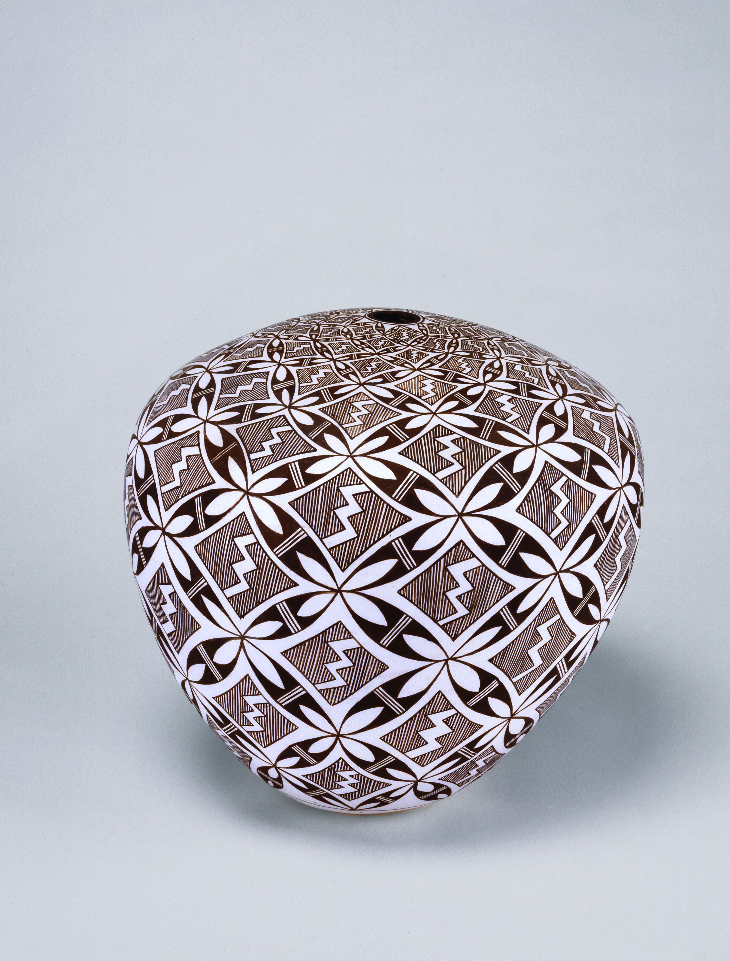 Round with a slight taper toward the base and a tiny opening at the top, this clay seed jar features a repeating pattern of abstracted flower shapes and a lightning bolt design. On the white surface the dark brown, delicately drawn designs expand and contract accordant with the form of the vessel.