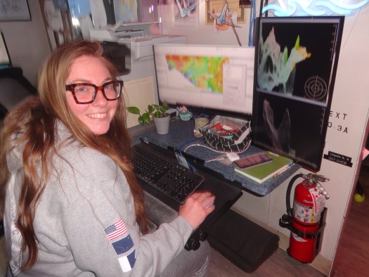 a young woman in a gray sweatshirt sits at a computer and smiles for the camera