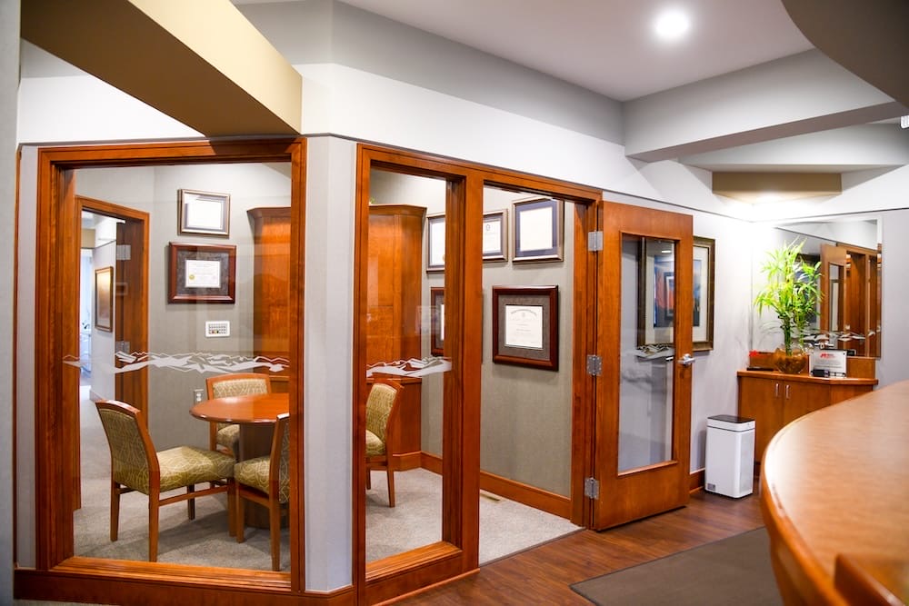 A modern Fort Collins office hallway features glass-walled rooms with wooden furniture, framed certificates on the walls, and plants on a counter. The space is brightly lit with recessed ceiling lights.