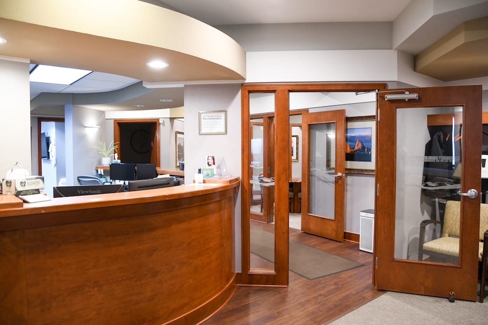 Reception area of a Fort Collins office with a curved wooden desk, computer monitor, and open wooden doors leading to rooms with chairs, framed art, and neutral-colored walls and flooring. The space is well-lit and tidy.