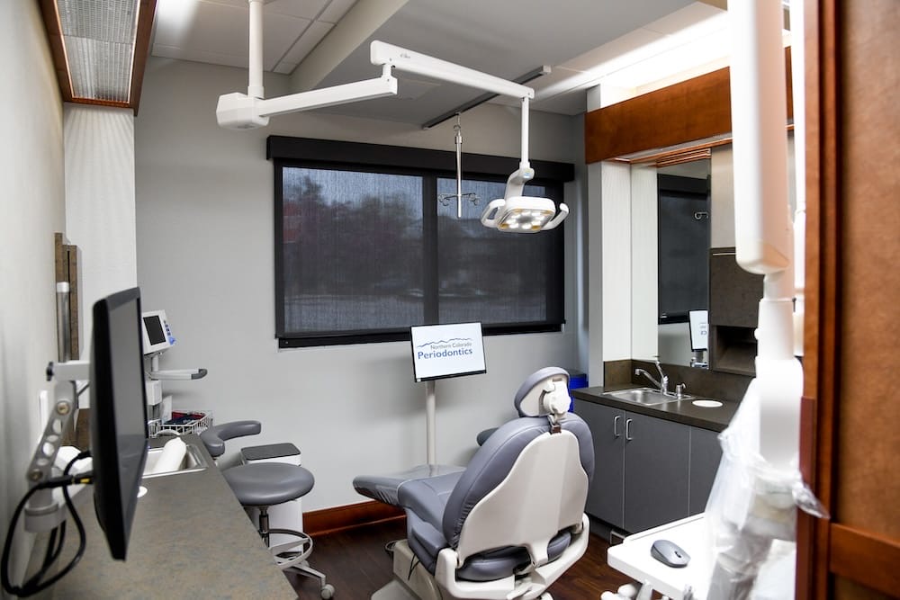 A modern Fort Collins dental office with an exam chair, overhead light, computer monitors, sink, cabinets, and dental equipment. A sign on a stand reads Periodontics. The room features large windows with shades.