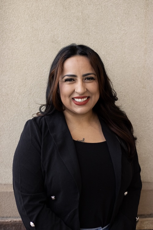 woman with tan skin and long dark hair, smiling and wearing a black shirt and blazer in front of a beige wall