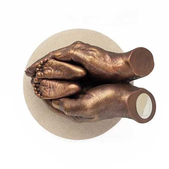 Escultura de resina de manos de adulto sosteniendo pies de bebé con acabado metálico en bronce. La pieza conmemorativa tiene una placa redonda plateada en el extremo y está montada sobre una base circular de color beige claro.