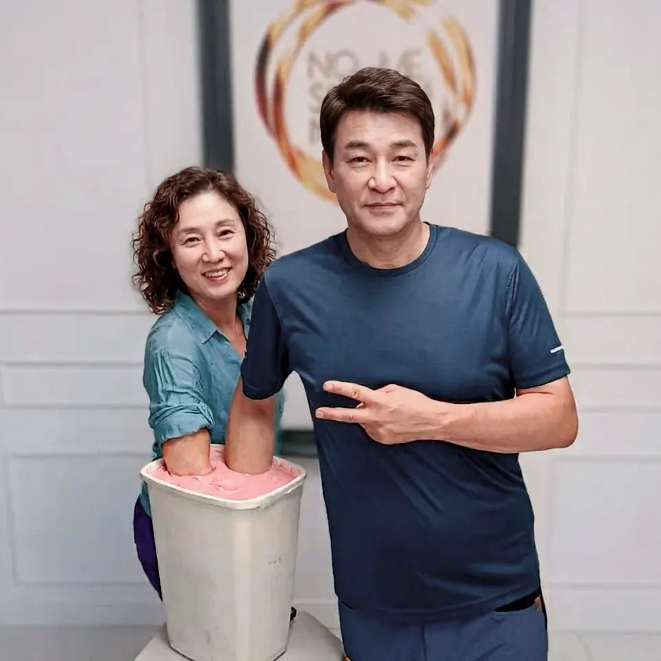 Una mujer sonriente y un hombre haciendo el signo de la paz introducen sus manos en un cubo rosa para crear un molde. Detrás, un cartel promocional de la experiencia "No me sueltes nunca" en Baños, La Rioja, vinculada al Camino de Santiago.