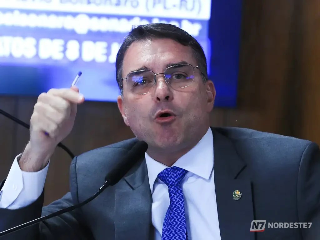 Foto em plano médio do senador Flávio Bolsonaro vestindo terno e gravata, com expressão séria durante uma sessão política.