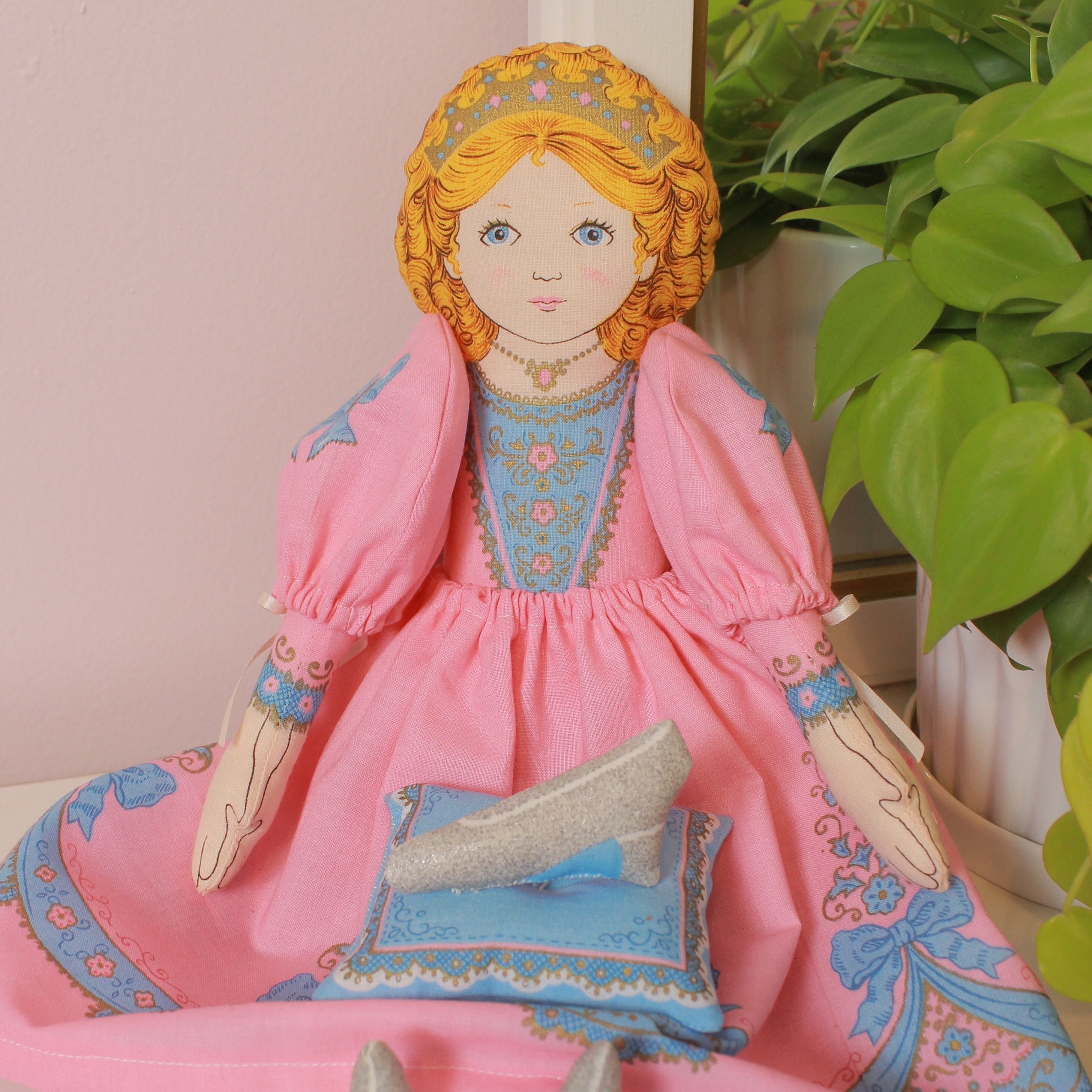 A fabric doll wearing a pink dress and holding a blue pillow with a glittery silver shoe on it.