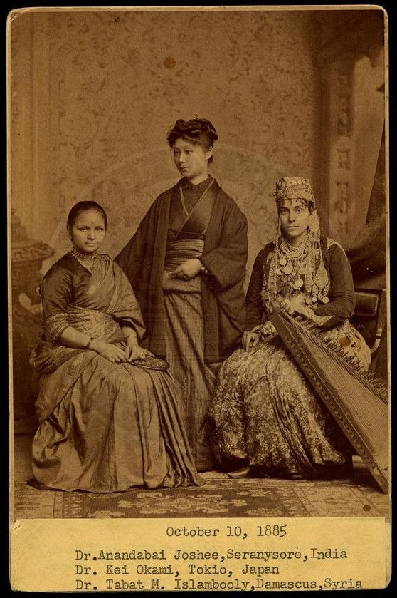 A photo of three female wearing traditional cloths, two sitting at the front and one standing behind