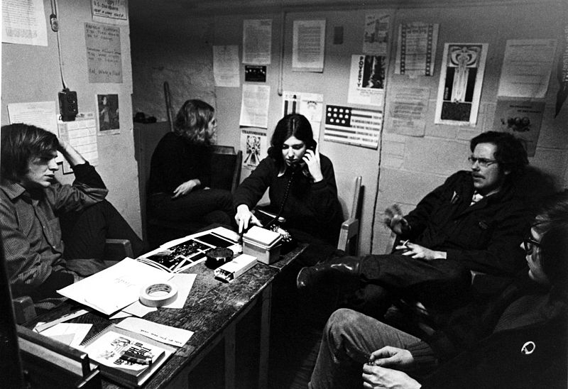 Black and white photograph of young people sitting around a table. One is on the phone, others are in conversation with each other.