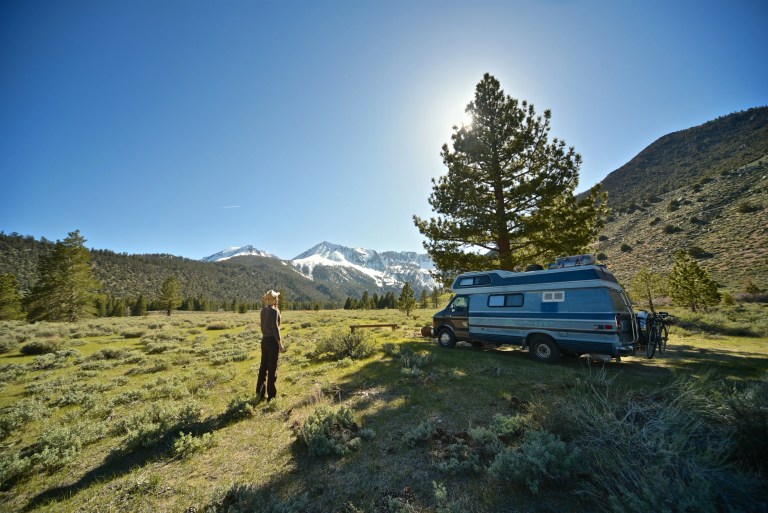 Femme regardant son van garé sous un sapin en plein milieu des montagnes.