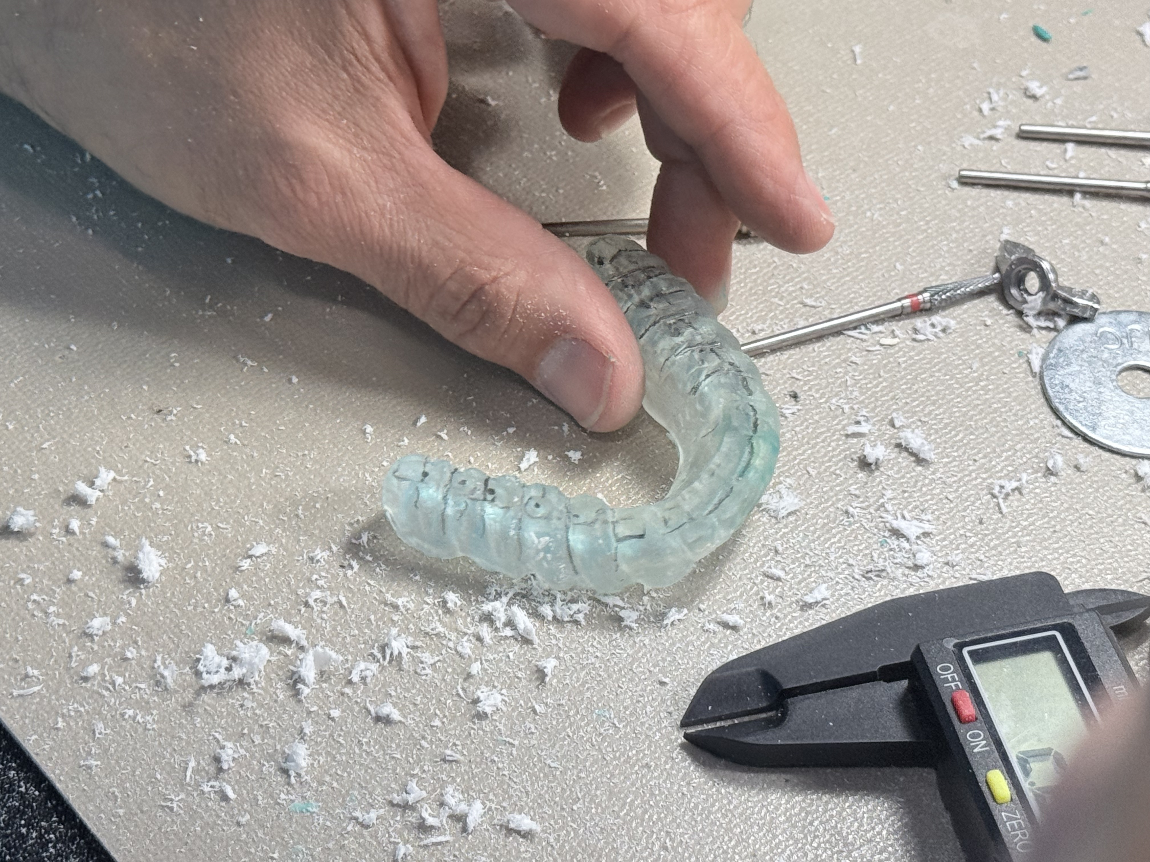 A clear GNM orthotic with pencil markings beside a digital caliper and acrylic shavings at the bench