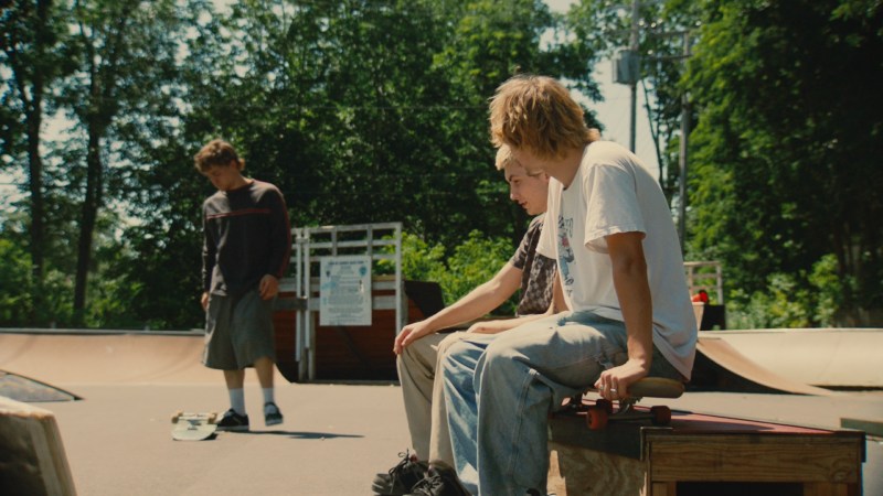 Two skateboarders sitting on a bench at a skate park, one with blonde hair wearing a white t-shirt, the other with brown hair in a black patterned shirt. A third skateboarder walks in the background with a skateboard.