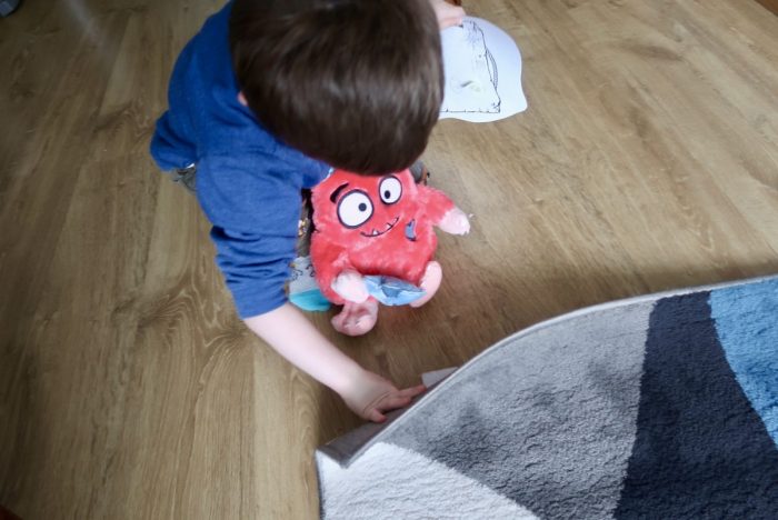 A little boy looking under a rug and holding a Love Monster stuffed toy