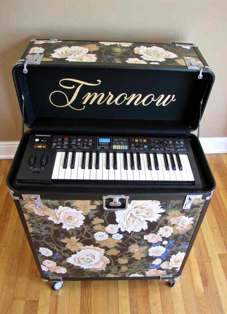 A keyboard synthesizer encased in a floral-patterned rolling case with large roses, featuring cursive text Tmronow inside the lid against a black background.