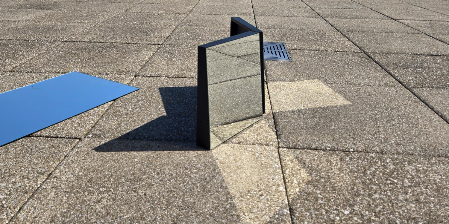 A horizontal, low-angle shot of two rectangular mirrors standing upright on a gray, tiled ground, forming a V-shape. Their dark frames cast long shadows on the ground. A third mirror is lying flat on the left side of the frame, reflecting a clear blue sky. A metal grate is visible in the background.