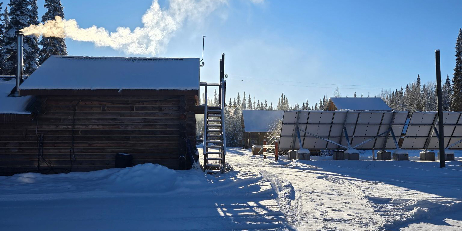 A horizontal, eye-level shot of a snow-covered, rustic log cabin on the left with smoke coming from its chimney. In the center, a staircase covered in snow leads to an upper deck. On the right, large solar panels are set up on the snow, angled toward the sun. In the background, there is a thick forest of snow-covered pine trees under a clear blue sky.