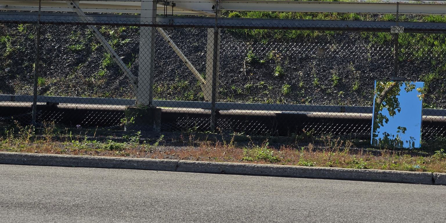 A long, horizontal, full-body shot of a large, rectangular mirror standing upright on a grassy embankment next to a paved road. The mirror is reflecting a clear blue sky and some tree leaves. A chain-link fence is visible behind the mirror, with a dirt slope and an industrial pipe in the background.